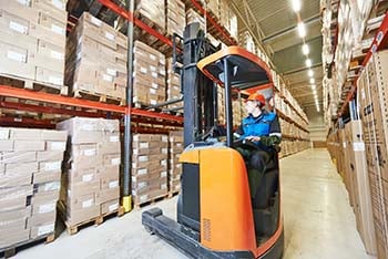 Alt text: "A warehouse worker operating an orange forklift in an aisle between tall racks filled with organized boxes and goods on pallets.