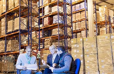 Three people, possibly a team of warehouse managers or workers, are seated and discussing over papers and a digital tablet in a large warehouse full of high shelving stocked with numerous boxes on pallets.