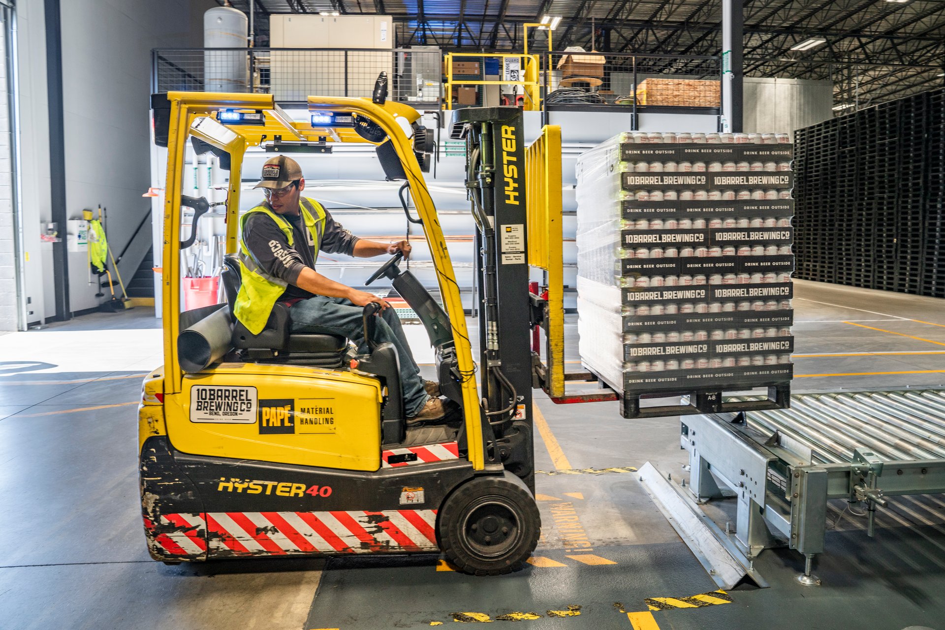 A worker in a high-visibility vest operates a yellow forklift inside a warehouse. The forklift is lifting a pallet stacked with boxes. The facility has a high ceiling, and industrial equipment and shelving can be seen in the background.