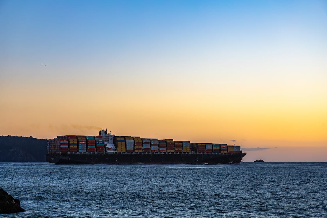 A large cargo ship laden with multicolored containers sails near the coastline during sunset, with the golden hues of the setting sun illuminating the sky and reflecting off the calm waters. In the distance, a silhouette of a hilly landscape can be seen.