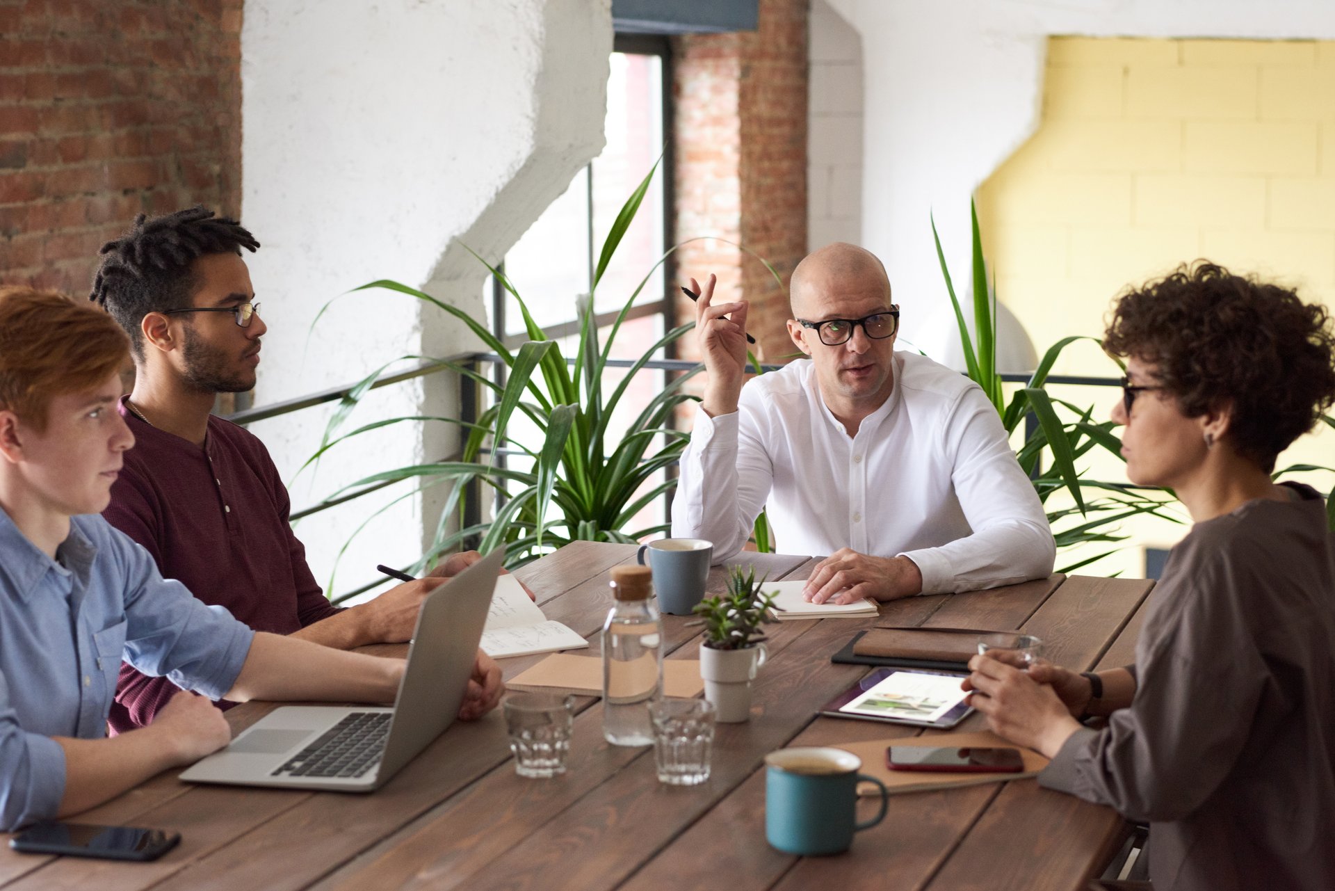 A group of four diverse individuals sit around a wooden table in a room with exposed brick walls and plants. They are engaged in a discussion, with one man gesturing with his hand. 
