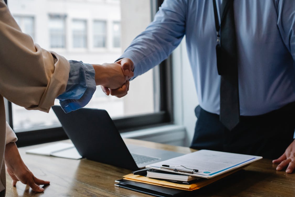 Two professionals shaking hands in an office setting, with a laptop, notepad, and other work essentials on the desk beside them. The backdrop features a window with a view of buildings outside. 